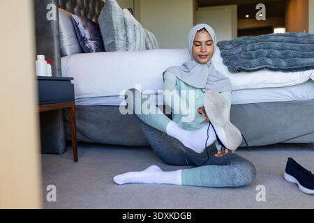Erwachsene Frau in grauem Hijab und mintgrünem Schnürschuh auf dem Schlafzimmerboden mit getuftetem Kopfteil Stockfoto