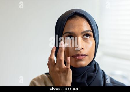 Afroamerikanische Frau glättet Augencreme mit Fingerspitzen im Zimmer in der Nähe von Fensterläden und trägt Hijab Stockfoto