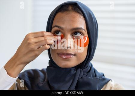 Afroamerikanische Frau im marineblauen Hijab, die orangen Hydrogel-Pflaster unter dem Auge an der Eitelkeit anpasst Stockfoto