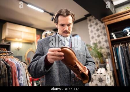 Ernsthaft gutaussehender Mann, der darüber nachdenkt, elegante Lederschuhe im Second Hand Vintage Shop zu kaufen. Stockfoto