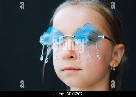 Nahaufnahme-Porträt eines ernsten Kindes mit lustiger blauer, wolkenförmiger Sonnenbrille mit Kristallregentropfen. Konzepte von verborgenen Emotionen, Traurigkeit, Fantasie Stockfoto