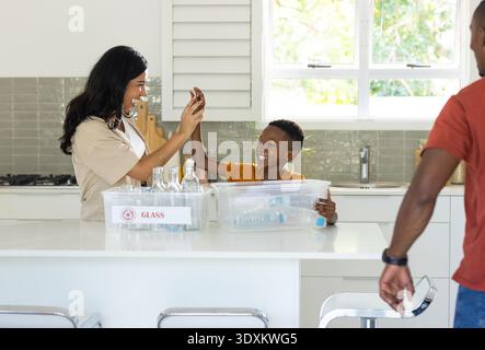 Vielfältige Familie mit Kinderhochfiving, Sortierung von Wertstoffen auf weißer Insel mit Mülleimern mit GLASMARKIERUNG Stockfoto
