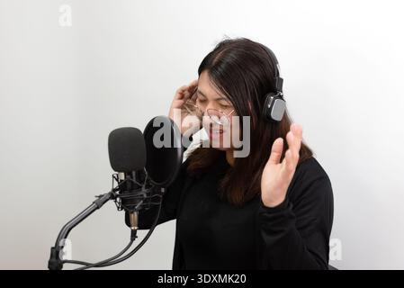 Junge Frau mit Kopfhörern, die beim Aufnehmen von Podcasts oder Musik in Studioumgebungen, die digitale Inhalte darstellen, in das Mikrofon singen oder sprechen Stockfoto