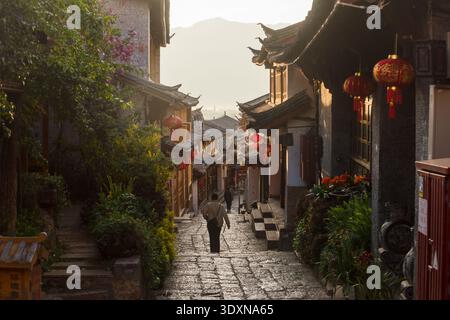 LIJIANG, YUNNAN, CHINA - 12. APRIL 2025: Eine enge gepflasterte Straße in der Altstadt von Lijiang während des Sonnenaufgangs, mit traditionellen Holzgebäuden Stockfoto