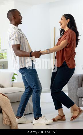 Hispanische Frau und afroamerikanischer Mann, der Hände hält und sich im Wohnzimmer in der Nähe des grauen Sofas bewegt Stockfoto