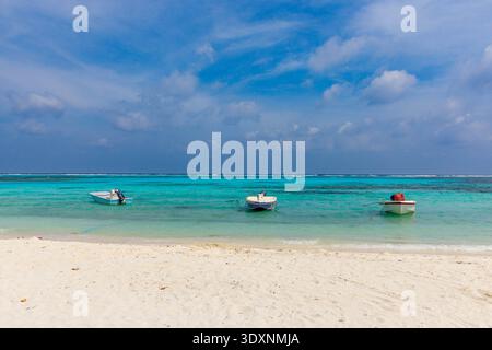 Türkisfarbenes Wasser trifft unter hellem Himmel auf weißen Sand. Drei kleine Boote schweben ruhig am Ufer. Eine Einzelfigur sitzt in einem und genießt den Frieden. Perfe Stockfoto