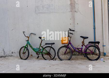 Zwei Motorräder lehnen sich gegen eine verwitterte Wand. Eine grüne, eine lila, beide zeigen Gebrauchsspuren. Der gelbe Korb verleiht dem lila Motorrad Farbe. Hinweis auf staubigen Boden Stockfoto