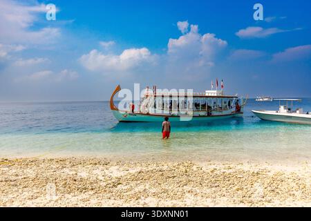 Türkisfarbenes Wasser trifft auf goldenen Sand unter hellblauem Himmel. Eine Einzelfigur schlendert barfuß in die Nähe sanfter Wellen. Weißes Boot mit Flagge schwimmt friedlich nebenan Stockfoto