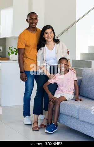 Vielfältige Familie lächelt, posiert im modernen Wohnbereich auf grauem Sofa in der Nähe der Küchenzeile Stockfoto