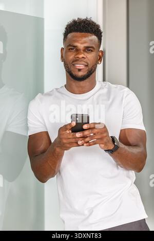 Ein Mann steht lässig vor einer schlanken Glaswand und konzentriert sich auf sein Smartphone. Er trägt ein einfarbiges weißes T-Shirt und hat einen selbstbewussten Ausdruck, der verkörpert Stockfoto