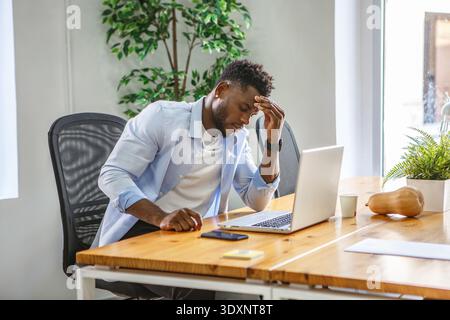 In einer modernen Büroumgebung wirkt ein Mann gestresst, während er an seinem Holztisch sitzt. Er hält seine Stirn, sichtbar besorgt, während er an seiner arbeitet Stockfoto