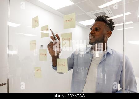 Ein Mann arbeitet aktiv an Brainstorming-Sitzungen und schreibt auf Haftnotizen, die an einer klaren Tafel angebracht sind, umgeben von einer modernen Büroumgebung Stockfoto