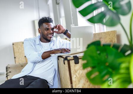 Ein junger Mann arbeitet an seinem Laptop in einer gut beleuchteten, modernen Büroumgebung und genießt einen entspannten Moment inmitten von Innenanlagen. Stockfoto