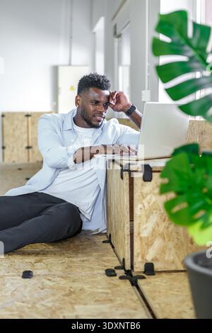 Ein junger Mann sitzt bequem auf dem Boden eines stilvollen Büros und benutzt einen Laptop für die Arbeit. Tageslicht erfüllt den Raum, und Grün sorgt für eine erfrischende Atmosphäre Stockfoto