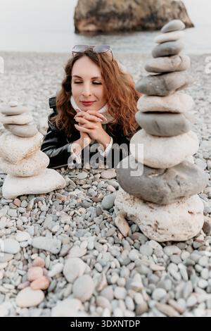 Frau mit Steinkieseln entspannt rothaarig liegend an einem felsigen Ufer mit gestapelten Steinen für eine meditative Zen-Atmosphäre Stockfoto