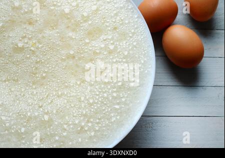 Eine weiße Schüssel mit geschlagenem Eiweiß, mit einer schaumigen Konsistenz und zahlreichen Luftblasen, auf grauem Holzhintergrund. Kulinarisches Kreativkonzept. Stockfoto