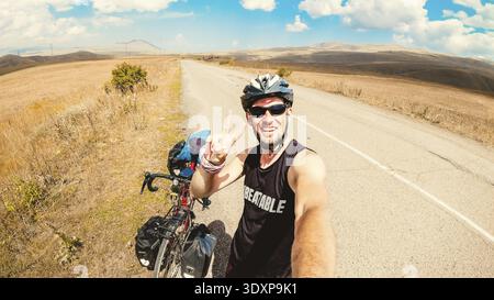 Ein weißer Mann steht am Fahrrad allein und fährt durch die ruhige Landschaft, voll mit Taschen im Freien, im Sommer zeigt die Hitze Daumen hoch. Kreative ec Stockfoto