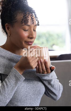 Afroamerikanerin, die einen Becher hält, der auf dem Sofa neben dem Fenster Aroma genießt und grauen Pullover und Ringe trägt Stockfoto
