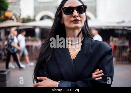 Stilvolle Frau mit langen dunklen Haaren und Sonnenbrille steht selbstbewusst in einer urbanen Umgebung, die Arme gekreuzt, Eleganz und moderne Mode in einer lebendigen Atmosphäre ausstrahlt || Model erscheint Stockfoto