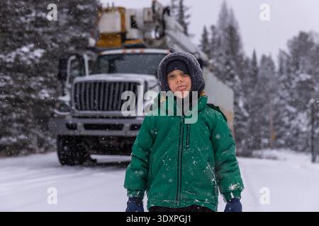 Kind, das während des Schneefalls die Zunge herausragt, mit einem Schaufelwagen im Hintergrund, in einem verschneiten Wald || Model veröffentlicht Stockfoto