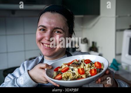 Porträt einer lächelnden Frau, die einen Teller Fusilli-Pasta mit Pilzen, geriebenem Käse und Kirschtomaten hält. Stockfoto