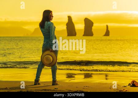 Eine Frau in Denim steht an einem Sandstrand mit Hut und beobachtet ferne Ozeanstapel zur goldenen Stunde. Das warme, gelbe Licht erzeugt eine ruhige, besinnliche Atmosphäre Stockfoto