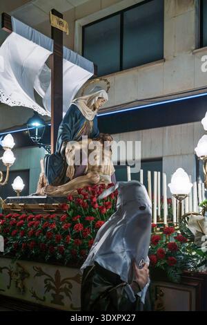 Karwochenprozession in Spanien mit einem religiösen Wagen Pietà, der mit roten Rosen und einem Buße in einer traditionellen weißen Kapuze geschmückt ist. Stockfoto