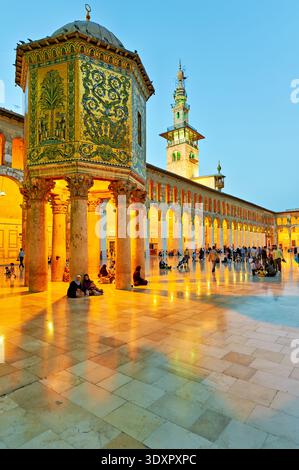 Besucher sitzen ruhig im Innenhof der Umayyaden-Moschee in Damaskus, während die Sonne untergeht und sanftes Licht über die historische Stätte zaubert. Stockfoto