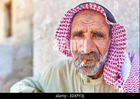 Ein älterer Mann sitzt in Aleppo, Syrien, trägt traditionelle Kleidung und einen rot-weißen Schal, während er eine Zigarette raucht. Stockfoto