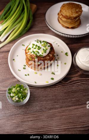 Gestapelte goldene Kartoffelpfannkuchen (Latkes) auf einer weißen Keramikplatte, garniert mit cremiger Sauerrahm und gehackten grünen Zwiebeln, auf einem rustikalen Holz angeordnet Stockfoto