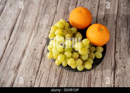 Nahaufnahme des Stilllebens eines Reifen grünen Traubenbusches neben zwei hellen Orangen auf einem dunklen Schieferteller auf einer rustikalen, verwitterten Holzplatte, warm gebadet Stockfoto