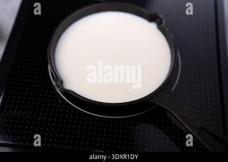 Nahaufnahme einer schwarzen gusseisernen Pfanne, gefüllt mit weißer Milch, auf einem glänzenden schwarzen Induktionskochfeld, einer kontraststarken Minimalküche Stockfoto