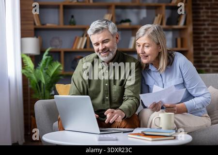 Lächelndes Seniorenpaar, das mit einem Laptop Ausgaben berechnet und Finanzdokumente in ihrem gemütlichen Wohnzimmer überprüft, um ihren Ruhestand und die Zukunft zu planen Stockfoto