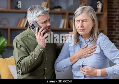 Besorgter Mann wählt Notdienste, während sein älterer Partner ihre Brust bei starken Schmerzen zu Hause, dringenden medizinischen Krisen und Unterstützung während der Notlage umklammert Stockfoto