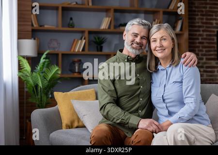 Reife Ehemann und Ehefrau sitzen auf einer modernen Wohnzimmercouch, lächelnd und entspannt und teilen einen warmen, komfortablen Moment der Gesellschaft, Liebe und Zufriedenheit zu Hause Stockfoto