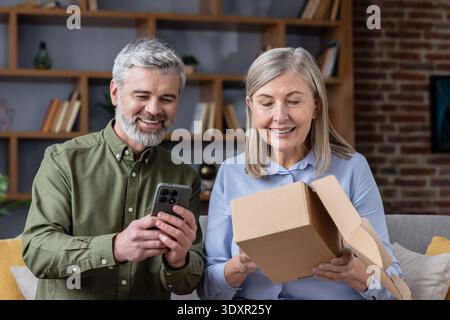 Glückliches Seniorenpaar auf einem Sofa, das eine Lieferung auspackt, eine Frau, die ein Papppaket öffnet, während der Mann ein Smartphone überprüft, entspanntes E-Commerce-Shopping und Komfort zu Hause genießt Stockfoto