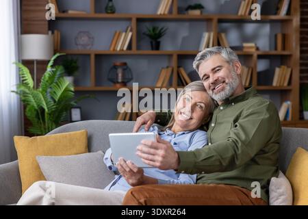 Glückliches Seniorenpaar, das sich auf dem Wohnzimmer-Sofa entspannt, lächelt und ein digitales Tablet teilt, während es zuhause surft, streamt und moderne Technologie genießt Stockfoto