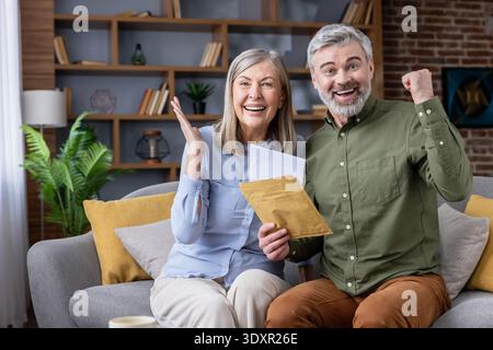Ein aufgeregtes Seniorenpaar sitzt auf einem Sofa, liest wichtige Post und jubelt mit fröhlichen Ausdrücken, feiert finanziellen Erfolg, eine Investitionsrendite oder eine Ruhestandsankündigung zu Hause Stockfoto