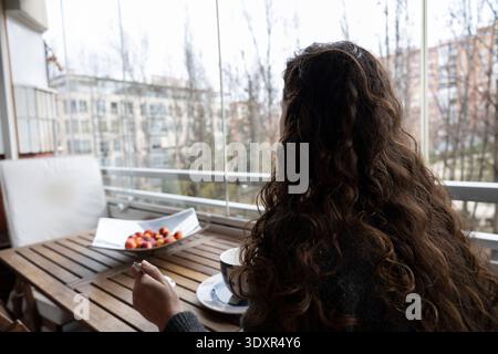 Frau, die an einem Holztisch auf einem Balkon sitzt und Kaffee und Obst trinkt und auf die Stadt blickt Stockfoto