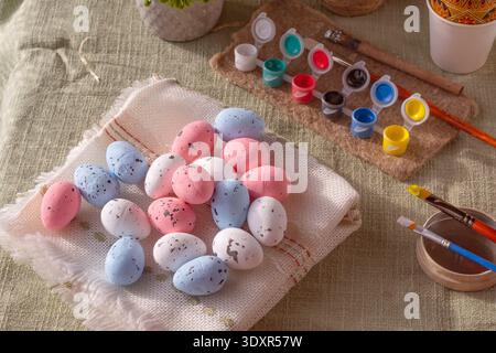 Pastellfarbene Ostereier mit Malutensilien und Pinseln auf rustikalem Textilhintergrund, Frühlingsferien-Kunsthandwerksszene Stockfoto