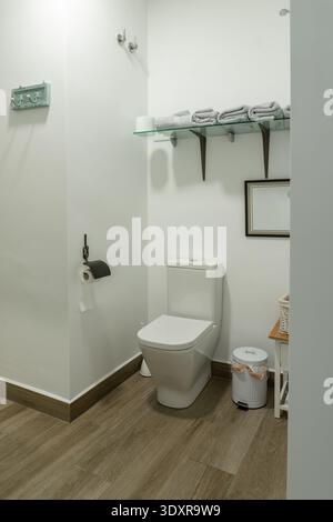 Modernes Badezimmer mit sauberer weißer Toilette, Glasablage mit Handtüchern, Holzboden und wichtigen Hygieneartikeln Stockfoto