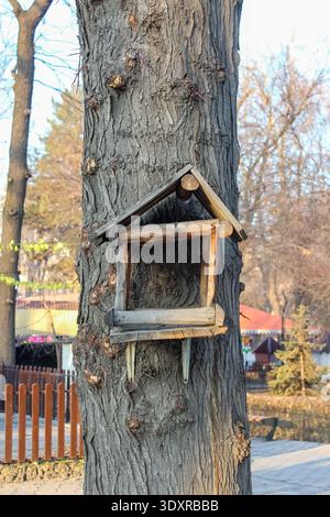 Eine rustikale, handgefertigte Vogelfutteranlage aus Holz befindet sich auf der strukturierten Rinde eines großen Baumes in einem sonnendurchfluteten Park oder Wald. Stockfoto