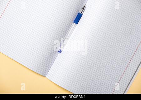 Ein offenes Notizbuch mit Graphpapier liegt auf einer gelben Oberfläche, begleitet von einem blauen Stift, der auf den Seiten liegt. Zurück zur Schule. Modelldesign Stockfoto