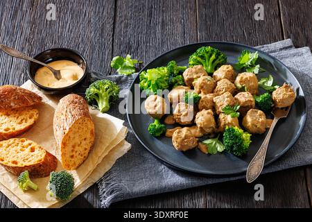 Belgische Gehaktballen, Fleischbällchen in cremebrauner Soße mit gedämpften Brokkoli-Röschen auf Teller mit Gabel, knuspriges glutenfreies Baguette auf dunklem Holztisch Stockfoto