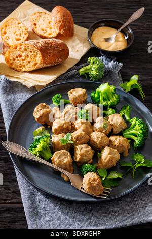 Belgische Gehaktballen, Fleischbällchen in cremebrauner Soße mit gedämpften Brokkoli-Röschen auf Teller mit Gabel, knuspriges glutenfreies Baguette auf dunklem Holztisch Stockfoto
