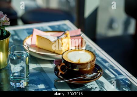 Kaffee mit Milchschaum und baskischer Käsekuchen auf der sonnigen Terrasse. Europäische Kaffeekultur, Frühstück, Dessert und Lifestyle-Konzept. Stockfoto