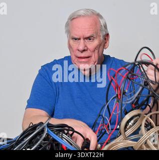 Ein älterer Mann in blauem Hemd siebt durch ein verworrenes Kabelgewirr auf dem Tisch mit Ausdruck der Frustration. Stockfoto