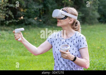 Eine fokussierte Frau verwendet VR-Brillen und -Controller, um an einer immersiven Lernsimulation teilzunehmen, während sie in der Natur sitzt. Stockfoto