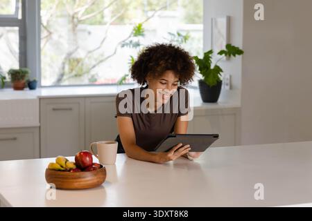 Afroamerikanische Frau mit braunem T-Stück, lehnt sich auf weiße Kücheninsel mit Tablette, Obst, Becher Stockfoto