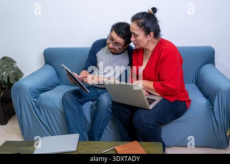 Ein älteres Ehepaar, das ein digitales Tablet und ein Notebook nutzt, um Online-Inhalte auf einem Sofa zu diskutieren Stockfoto
